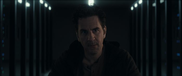 A man stands facing the camera in a dark server corridor, lit from directly above.